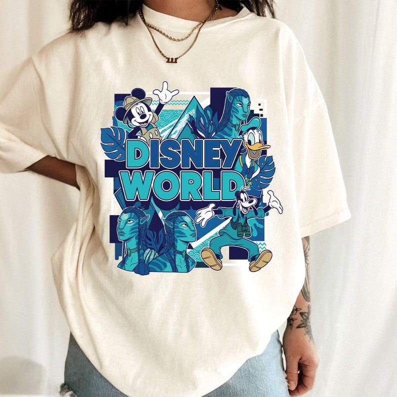 Puede incluir: Camiseta color crema con un dise&ntilde;o gr&aacute;fico. El dise&ntilde;o presenta el texto "DISNEY WORLD" en turquesa y azul, junto con ilustraciones de Mickey Mouse, el Pato Donald, Goofy y personajes de Avatar.