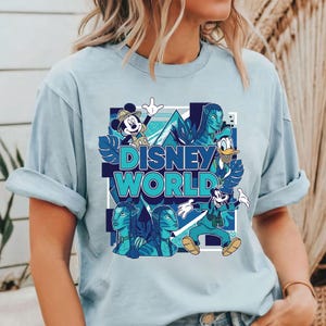 Puede incluir: Camiseta azul claro con un gr&aacute;fico de Disney World. El dise&ntilde;o incluye a Mickey Mouse, el Pato Donald, Goofy y personajes de Avatar. Las palabras "Disney World" est&aacute;n en turquesa, dentro de un cuadrado azul.
