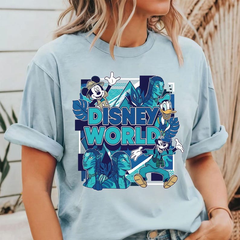 Puede incluir: Camiseta azul claro con un gr&aacute;fico de Disney World. El dise&ntilde;o incluye a Mickey Mouse, el Pato Donald, Goofy y personajes de Avatar. Las palabras "Disney World" est&aacute;n en turquesa, dentro de un cuadrado azul.
