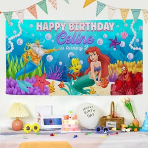 May include: A rectangular birthday banner featuring a blue underwater scene with King Triton, Ariel, Flounder, and Sebastian. The text reads HAPPY BIRTHDAY Celine is turning 2. The banner is decorated with bubbles, coral, and starfish, and is topped with colorful triangular bunting flags.