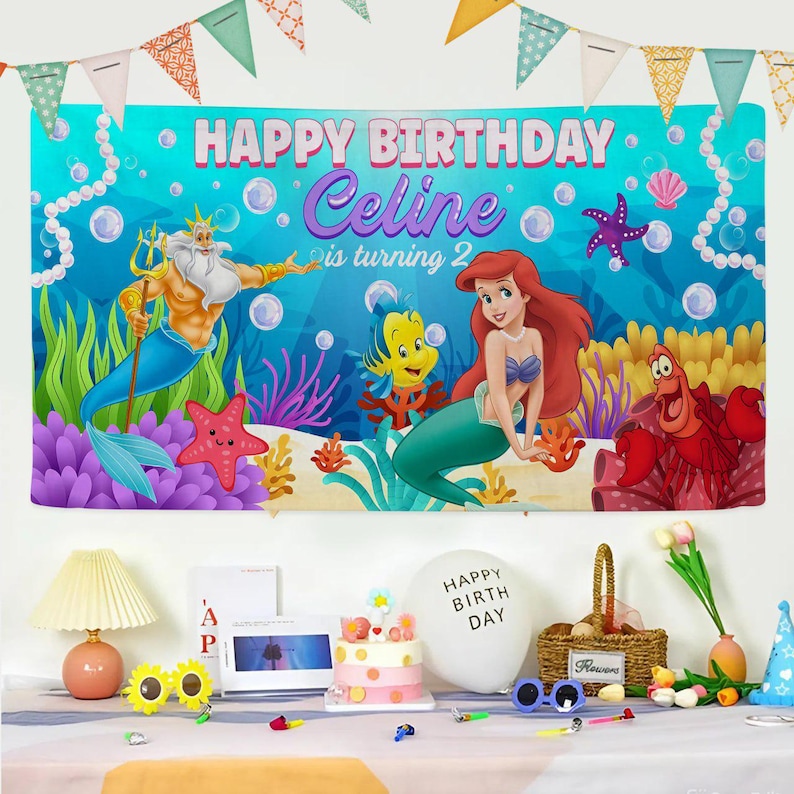 May include: A rectangular birthday banner featuring a blue underwater scene with King Triton, Ariel, Flounder, and Sebastian. The text reads HAPPY BIRTHDAY Celine is turning 2. The banner is decorated with bubbles, coral, and starfish, and is topped with colorful triangular bunting flags.
