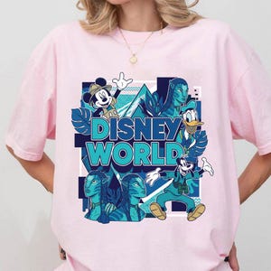 Puede incluir: Camiseta rosa claro con un gr&aacute;fico colorido de Disney World. El dise&ntilde;o incluye a Mickey Mouse, el Pato Donald, Goofy y personajes de Avatar, con las palabras "Disney World" en turquesa. Un dise&ntilde;o divertido y llamativo.