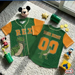 May include: Two green and orange baseball jerseys with dinosaur graphics. One jersey says "REX" and the other says "YOUR NAME" with the number "00". The jerseys are on a white rug with various toys.