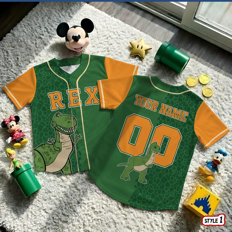 May include: Two green and orange baseball jerseys with dinosaur graphics. One jersey says "REX" and the other says "YOUR NAME" with the number "00". The jerseys are on a white rug with various toys.