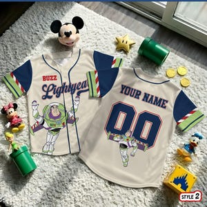 May include: Two beige baseball jerseys with navy blue sleeves and Buzz Lightyear graphics. One jersey says "Buzz Lightyear" and the other says "Your Name" with the number "00". The jerseys are surrounded by Disney-themed toys.
