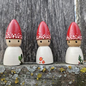 May include: Three wooden gnome figurines with red mushroom-shaped caps dotted with white spots. The gnomes have white bodies with painted details of flowers and greenery. They are displayed on a weathered wooden surface.