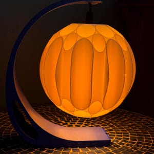 May include: A modern table lamp with a spherical, orange-yellow shade featuring a pattern of elongated ovals. The shade is suspended by a curved, dark blue frame. The lamp sits on a mosaic tile surface, creating a warm, ambient glow.