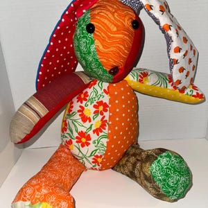 May include: A handmade patchwork bunny rabbit plush toy. The bunny features a variety of colorful fabrics, including red polka dots, orange floral patterns, and green rose prints. The bunny has black button eyes and a brown nose.