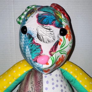 May include: A handmade fabric doll with a patchwork design. The doll's face features a mix of floral and patterned fabrics, with large black button eyes and a pink nose. The arms are yellow with white polka dots and green trim.