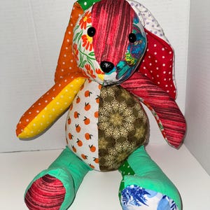 May include: A handmade patchwork bunny rabbit with a variety of colorful fabric patterns. The bunny features orange polka dots, floral prints, and geometric designs. It has black button eyes and a black nose. The bunny's ears are long and floppy, and its body is soft and plush.