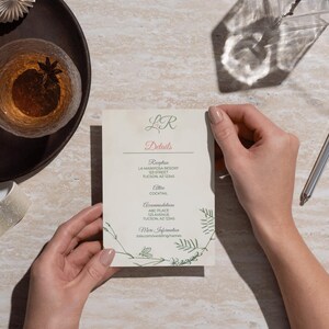 May include: A cream-colored wedding details card held by hands. The card features the initials "LR" in green, the word "Details" in pink, and event information in a serif font. Green botanical illustrations frame the text. A glass of amber liquid and a pen are in the background.