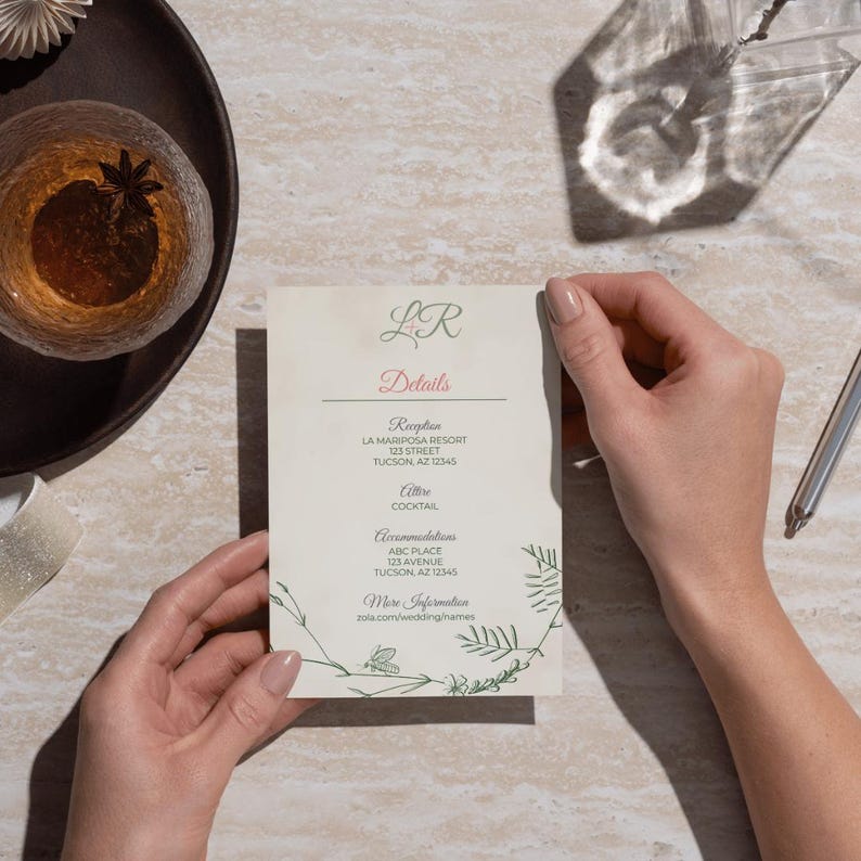 May include: A cream-colored wedding details card held by hands. The card features the initials "LR" in green, the word "Details" in pink, and event information in a serif font. Green botanical illustrations frame the text. A glass of amber liquid and a pen are in the background.