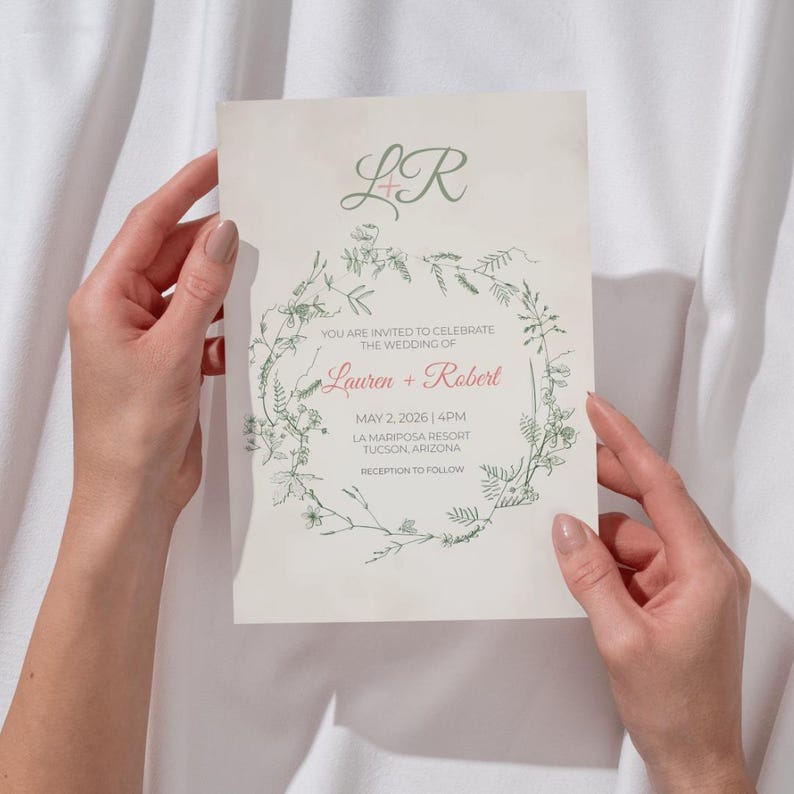 May include: A wedding invitation with a floral wreath design. The invitation features the names "Lauren + Robert" and event details. The text is in a mix of green and red fonts. The invitation is held against a white background.