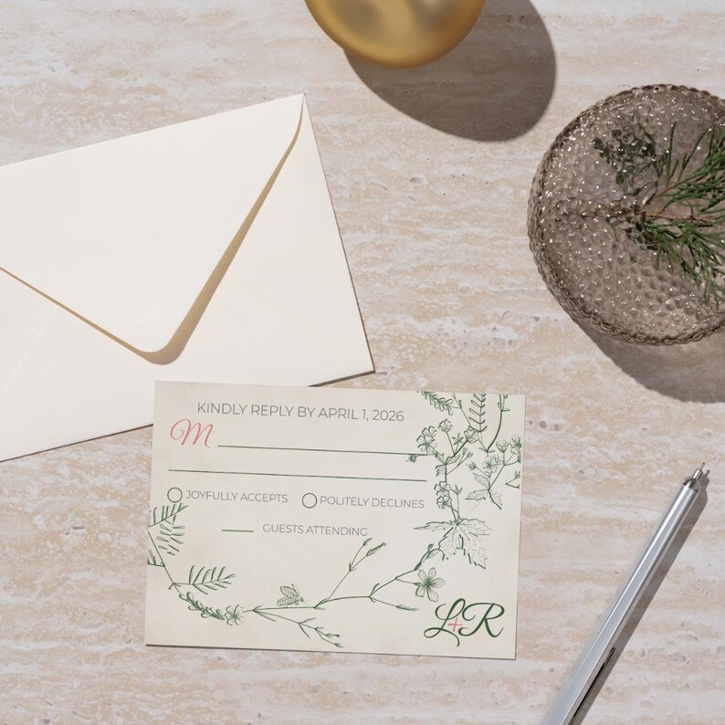 May include: A wedding response card with floral design and the text "KINDLY REPLY BY APRIL 1, 2026". The card includes options to accept or decline, with space for guest count. A matching envelope and pen are also present.