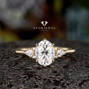 May include: A gold ring featuring a large oval-cut center stone flanked by two smaller triangle-cut side stones and tiny round accent stones. The text ZFORJEWEL JEWELRY is visible above the ring. The band is smooth and polished yellow gold.