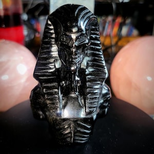 Puede incluir: Una escultura negra de un busto de faraón, con detalles intrincados y un acabado brillante. El busto se encuentra frente a un fondo borroso con esferas rosas y otros objetos. Es un artículo decorativo.