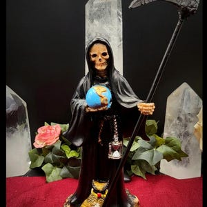 May include: A Grim Reaper figurine in a black robe, holding a blue globe and a scythe. The base is adorned with skulls. A small hourglass hangs from the Reaper's hand. The background includes quartz crystals and a rose.