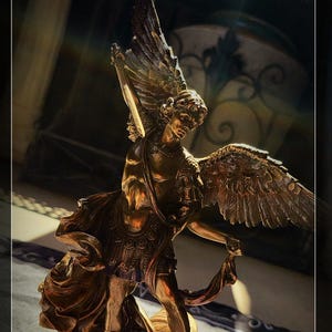 May include: A detailed bronze statue of an angel with large, detailed wings, holding a sword and a cloth. The angel is standing on a defeated figure. The statue has a golden hue and is set against a dark background.