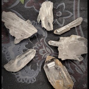 May include: A collection of clear quartz crystals of varying shapes and sizes. The crystals are arranged on a dark patterned surface, with some appearing translucent and others with a slight amber hue. The crystals are a natural, raw form.