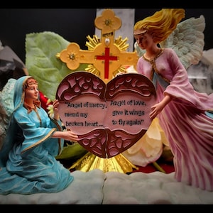 Op de afbeelding: Religieuze beeldjesset met twee engelenbeelden die een hartvormige plaquette met tekst vasthouden. Eén engel draagt een blauwe jurk, de ander roze. Op de achtergrond staat een gouden kruis.
