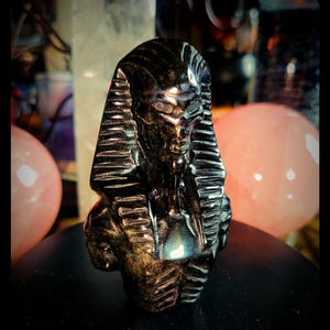 Può includere: Una scultura nera di un busto di faraone, con un copricapo e lineamenti del viso dettagliati. La scultura è ambientata su uno sfondo sfocato con sfere rosa. Il busto è su una superficie nera.