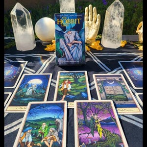 May include: A tarot card reading setup featuring "The Hobbit Tarot" deck. The image displays several tarot cards with colorful illustrations, quartz crystals, and a decorative hand. The deck box shows Gandalf. The background is a dark cloth.