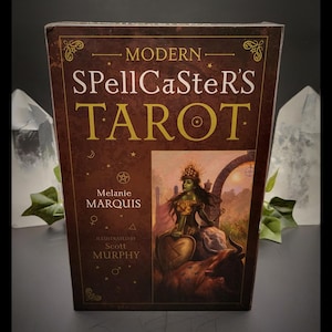 May include: A Modern SpellCaster's Tarot deck box. The box is brown with gold lettering and illustrations. The front features the title, author, and illustrator's names, along with a fantasy illustration of a woman with a shield.