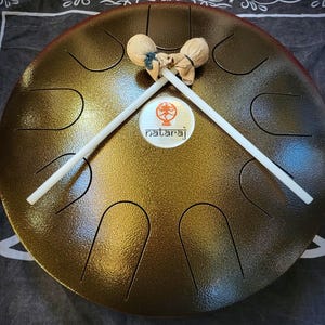 May include: A bronze-colored steel tongue drum with a circular shape and a textured surface. The drum has several curved slots cut into the top. Two white drumsticks with soft, rounded heads rest on the drum. A circular logo with the word "nataraj" is in the center.