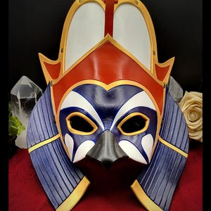 Può includere: Una maschera artigianale con un design a forma di uccello, con schema di colori rosso, bianco, blu e oro. La maschera ha un becco prominente e ali stilizzate, con dettagli intricati e una consistenza simile alla pelle. Per costumi o decorazioni.