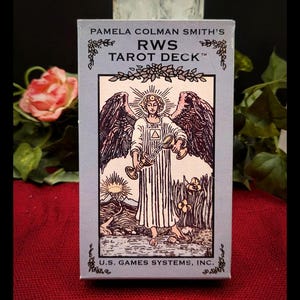 May include: A tarot deck box with the text "PAMELA COLMAN SMITH'S RWS TAROT DECK" and an illustration of an angel. The box is light blue with black and white artwork. The deck is from U.S. GAMES SYSTEMS, INC.
