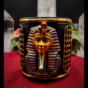 May include: A white ceramic mug with a black handle and the text "But First, Coffee" in a bold, black font. The mug is set against a blurred background, suggesting a cosy setting.