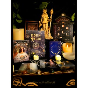 Puede incluir: Una colección de objetos místicos, incluyendo libros titulados "Moon Magic" y "Queen of the Moon Oracle", velas y una estatua de una mujer con un bastón. Un letrero dice "Magic in all of us".