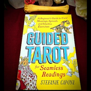 May include: A yellow book titled "GUIDED TAROT" with the subtitle "A Beginner's Guide to Card Meanings, Spreads, and Intuitive Exercises." The cover features illustrations of angels and a woman in a robe. A bone is placed above the book.