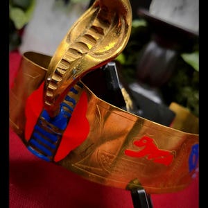 Puede incluir: Una pulsera dorada con un diseño de cabeza de cobra. La pulsera presenta detalles en rojo y azul y una figura roja estilizada. La pulsera se exhibe sobre una superficie roja.