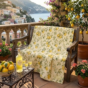Puede incluir: Un banco de madera con una colcha crema estampada con limones y hojas. Una botella de Limoncello y copas están sobre una mesa de hierro forjado. También se ve un cuenco con limones y un limonero.