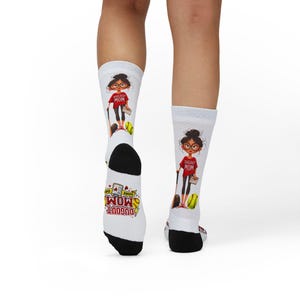 May include: White socks with a black heel and toe, featuring a cartoon illustration of a woman in a red shirt that says "DUGOUT MOM". The socks also have a baseball and the words "GAME DAY".
