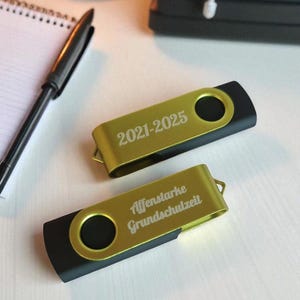 Könnte beinhalten: Zwei gold-schwarze USB-Sticks. Auf einem Stick steht "2021-2025". Auf dem anderen Stick steht "Affenstarke Grundschulzeit". Ein schwarzer Stift und ein Notizblock sind im Hintergrund.