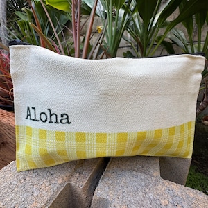 May include: Cream-colored canvas pouch with a yellow gingham bottom section. The word "Aloha" is printed in black on the upper portion. The pouch has a black zipper and is resting on a stone surface.