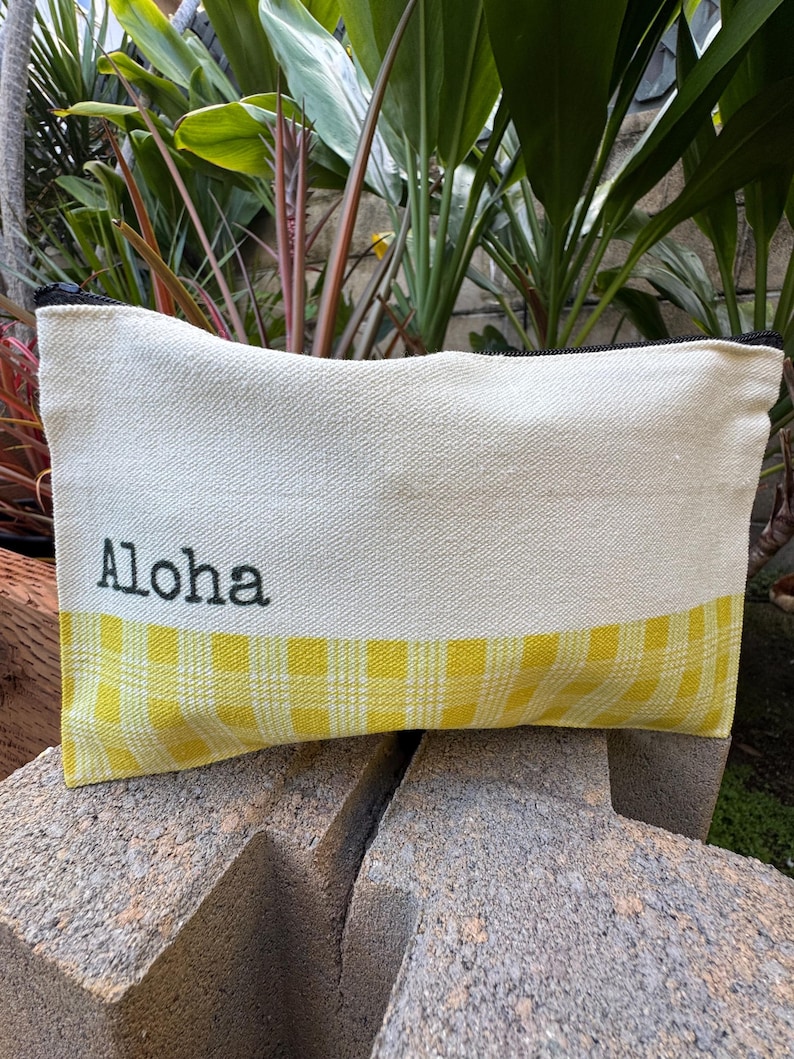 May include: Cream-colored canvas pouch with a yellow gingham bottom section. The word "Aloha" is printed in black on the upper portion. The pouch has a black zipper and is resting on a stone surface.