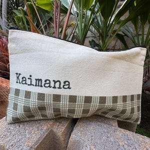 May include: Cream-colored canvas pouch with a black zipper. The bottom half features a green and brown plaid pattern. The word "Kaimana" is printed in black letters across the top.