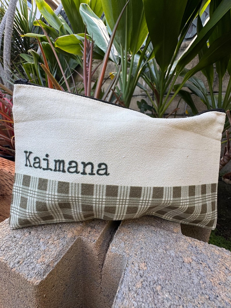 May include: Cream-colored canvas pouch with a black zipper. The bottom half features a green and brown plaid pattern. The word "Kaimana" is printed in black letters across the top.