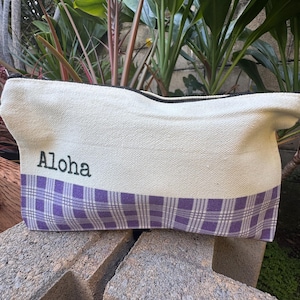 May include: Cream-colored canvas bag with a purple plaid pattern at the bottom. The word "Aloha" is printed in black on the upper portion of the bag. The bag has a black zipper and is sitting on a gray stone surface.
