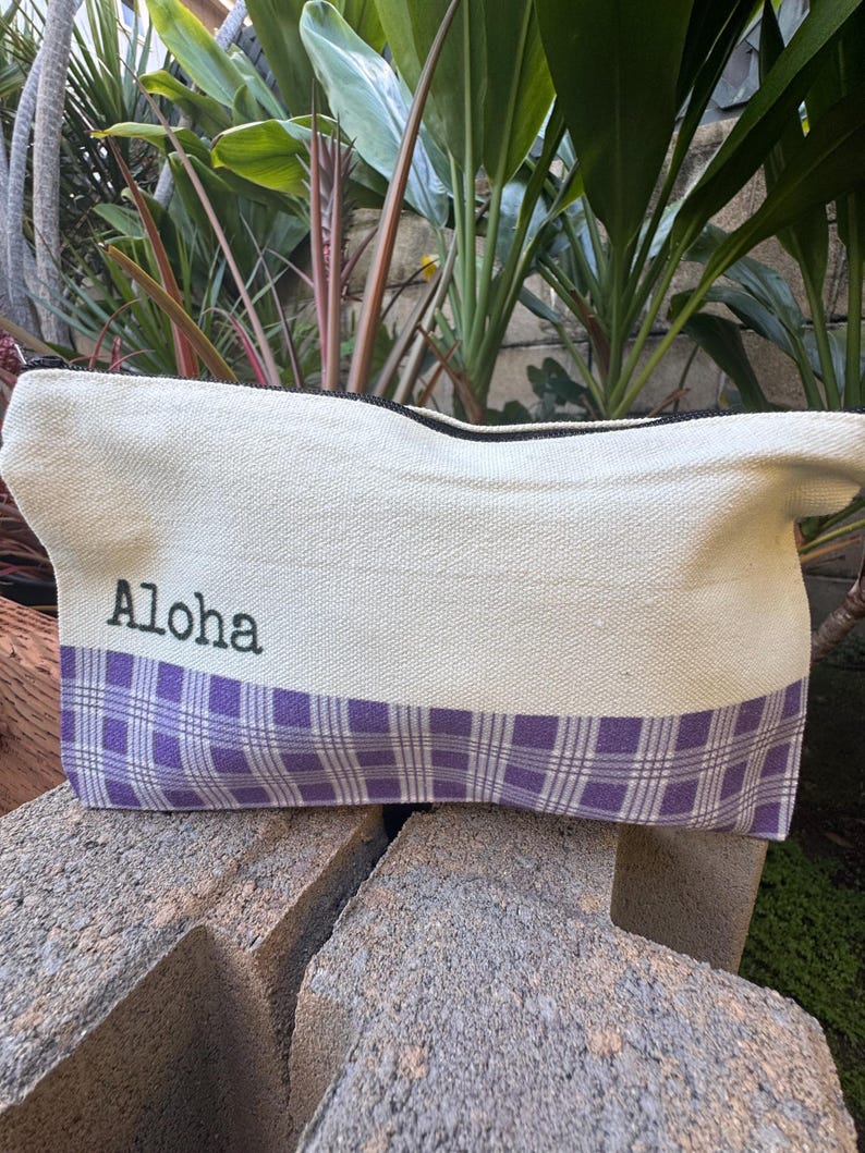 May include: Cream-colored canvas bag with a purple plaid pattern at the bottom. The word "Aloha" is printed in black on the upper portion of the bag. The bag has a black zipper and is sitting on a gray stone surface.
