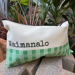 May include: Rectangular canvas pouch with a black zipper and the word "Waimanalo" printed in black. The bottom section features a green and white plaid pattern. The pouch is resting on a rough, gray surface.