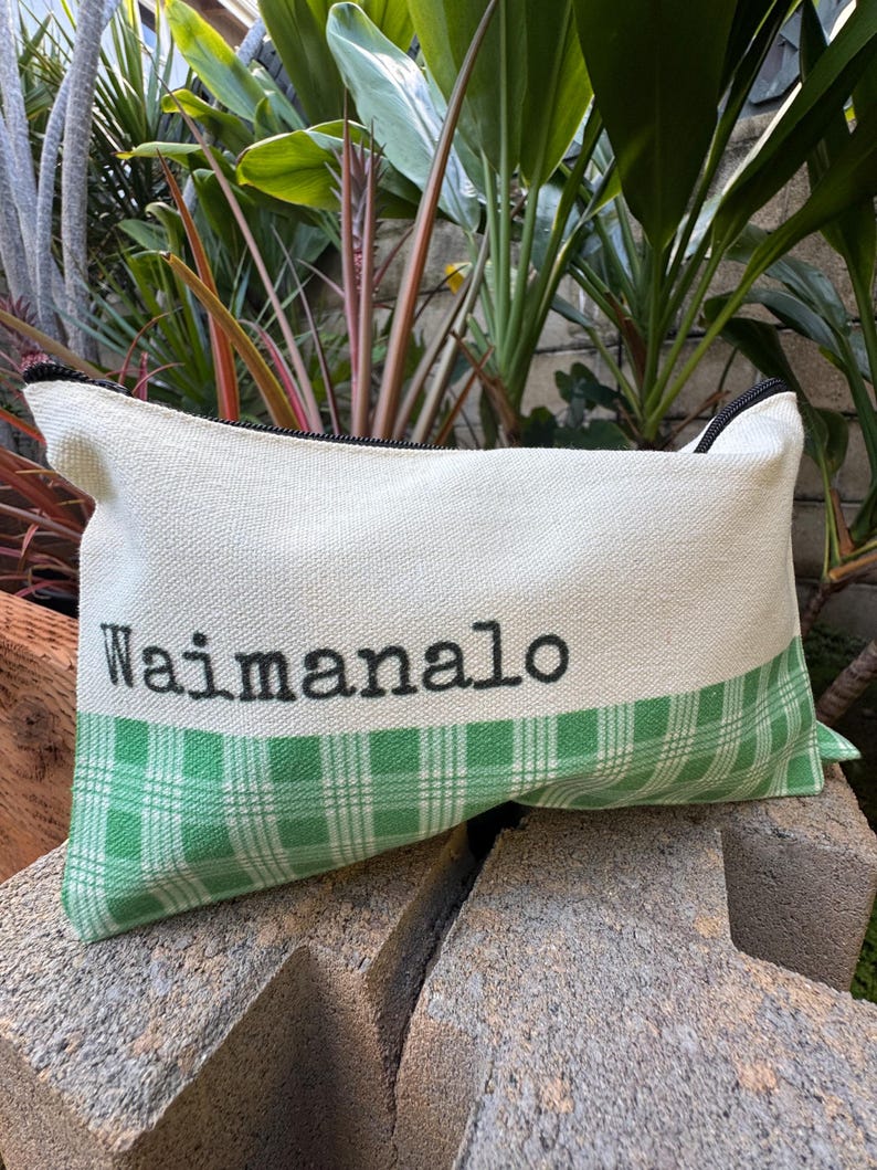 May include: Rectangular canvas pouch with a black zipper and the word "Waimanalo" printed in black. The bottom section features a green and white plaid pattern. The pouch is resting on a rough, gray surface.