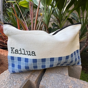 May include: A rectangular canvas pouch with a black zipper and the word "Kailua" embroidered in black. The bottom half features a blue and white plaid pattern. The pouch is resting on a concrete surface, with green foliage in the background.