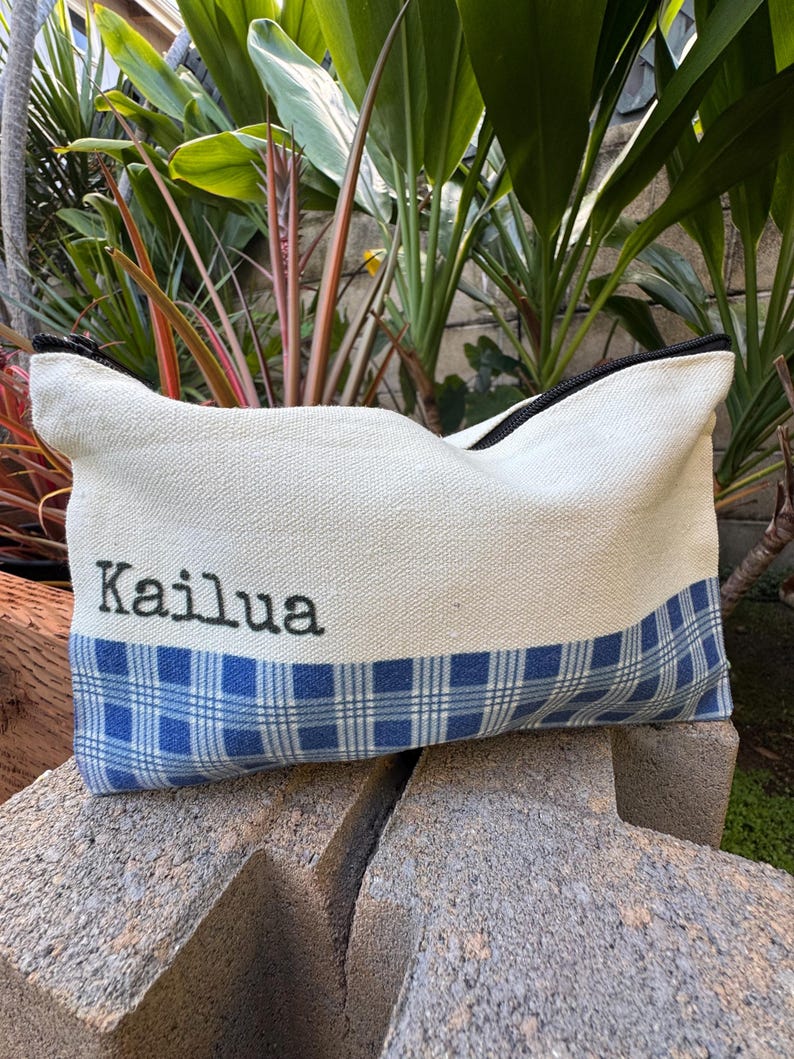 May include: A rectangular canvas pouch with a black zipper and the word "Kailua" embroidered in black. The bottom half features a blue and white plaid pattern. The pouch is resting on a concrete surface, with green foliage in the background.
