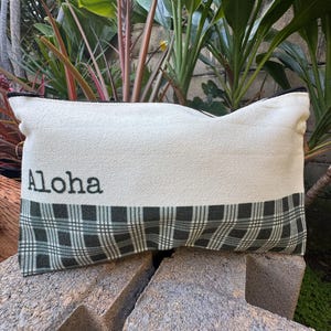 May include: Rectangular pillow with the word "Aloha" printed in black on a cream-colored fabric. The bottom section features a green and black plaid pattern. The pillow has a black trim.