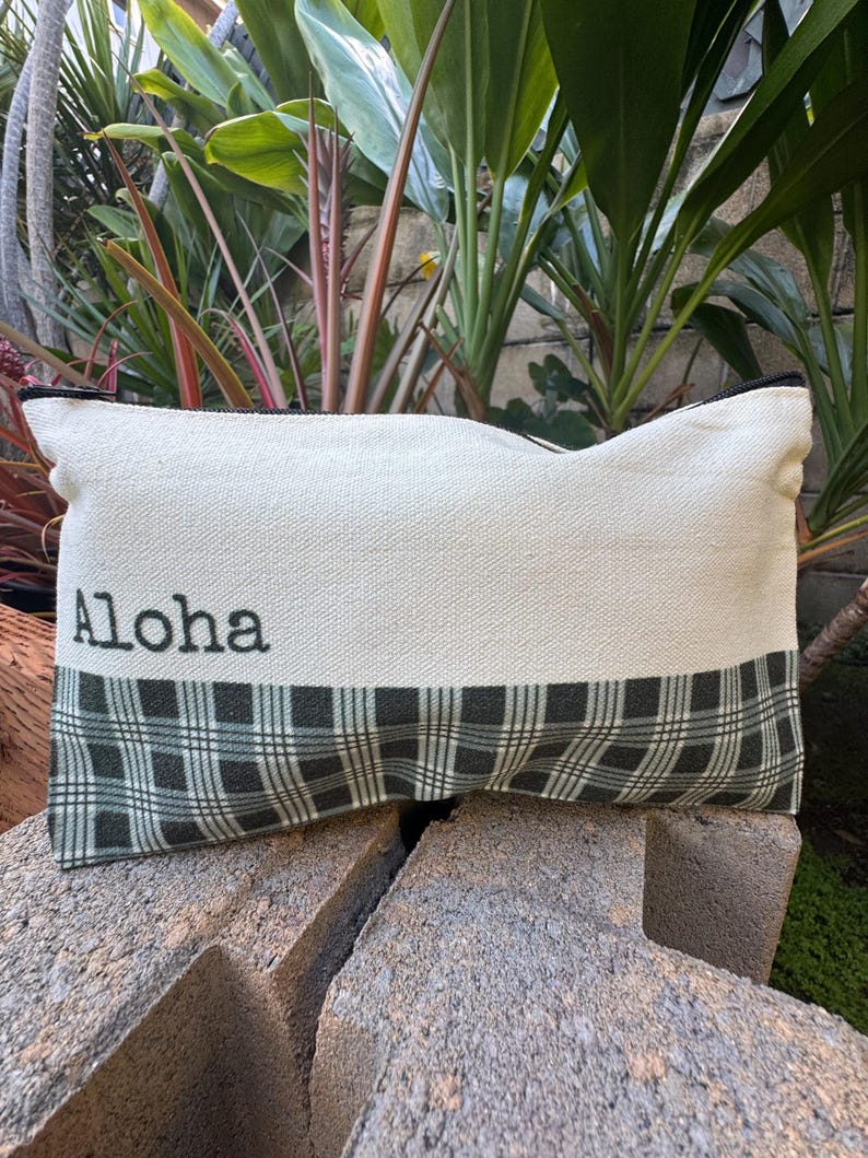 May include: Rectangular pillow with the word "Aloha" printed in black on a cream-colored fabric. The bottom section features a green and black plaid pattern. The pillow has a black trim.