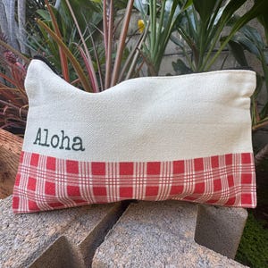 May include: Cream-colored rectangular pouch with a red and white plaid pattern at the bottom. The word "Aloha" is printed in black on the upper portion of the pouch. The pouch has a black zipper.